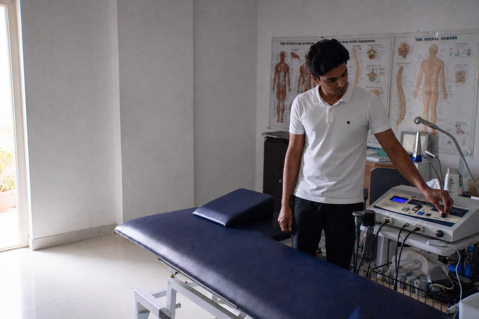 A physiotherapist setting the dial on an ultrasound therapy unit beside an empty treatment couch in a bright Hyderabad clinic room.