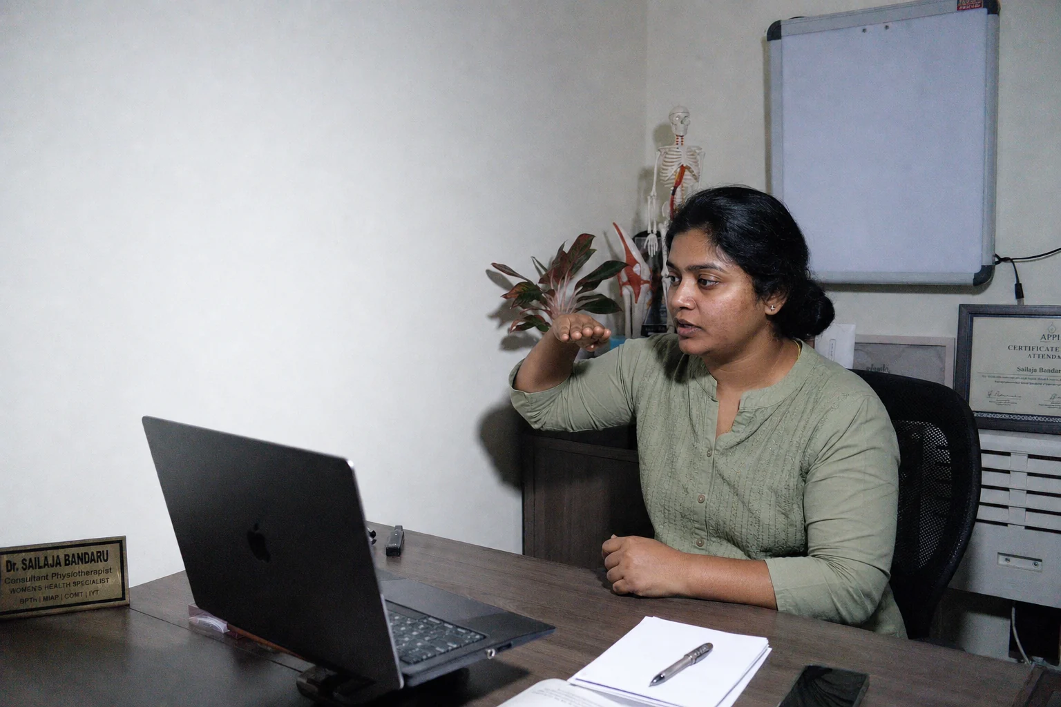 A physiotherapist at a clinic desk demonstrating a movement to a patient on her laptop screen.